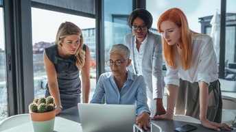 An einem Tisch sitzt eine Person mit kurzen, grauen Haare und Brille, die vor sich einen aufgeklappten Laptop stehen hat. Neben ihr stehen drei weitere Personen, die alle auf den Bildschirm des Laptops blicken