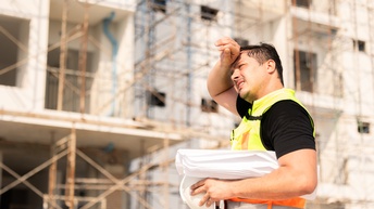 Seitliche Aufnahme einer Person mit Schutzweste, Helm und Papierrolle, die schwitzt und die rechte Hand auf die Stirn hält. Im Hintergrund ist eine Baustelle eines Gebäudes mit einem Gerüst