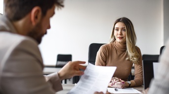 Eine Person blickt auf die Bewerbungsunterlagen einer zum Bewerbungsgespräch eingeladenen Person, die gegenüber von ihr sitzt.