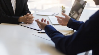 Zwei Personen sitzen sich an einem Tisch gegenüber. Vor der linken Person liegen Zettel. Auf dem Tisch sind außerdem ein Notizbuch und ein Laptop