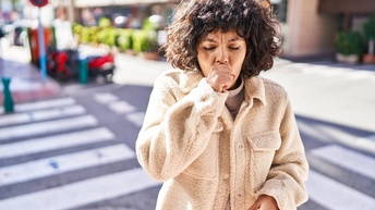 Frontalaufnahme einer Person in einem hellen Mantel und kinnlangen, dunklen Haaren. Die Person hält sich ihre rechte Faust vor den Mund, hustet hinein und hat die Augen geschlossen. Im Hintergrund sind zwei aufeinander treffende Straßen sowie ein Gebäude.