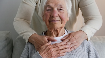 Nahaufnahme des Kopfes und des Brustbereichs einer sitzenden Person mit kurzen, grauen Haaren. Hinter dieser Person steht eine weitere Person, die ihre Hände auf die vordere Person legt. Die sitzende Person hat ihre rechte Hand auf der anderen Person.