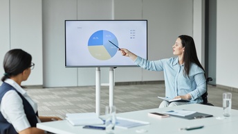 Person im Rollstuhl sitzt vor einem Monitor und zeigt mit einem Stift auf ein Diagramm während eine andere Person am Besprechungstisch sitzt