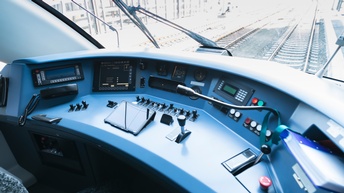 Hinter einer Glasscheibe verlaufen Gleise. Vor der Glasscheibe ist ein großer, blauer Tisch, der mit der Glasscheibe teilweise verbunden ist. Auf dem Tisch sind verschiedene Knöpfe, Schalter, ein Mikrofon, Hebel, ein Telefon, Bildschirme und ein Tablet