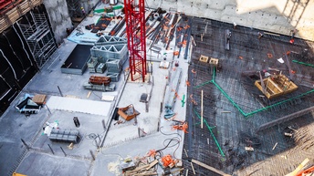 Baustelle in Vogelperspektive: Betonierte und mit Stahlkonstrukten gebauter Untergrund, darauf Maschinen, Baustoffe und Kran stehend