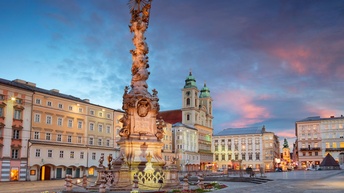 Die Pestsäule in Linz auf dem Hauptplatz bei Abenddämmerung. Der Platz wird von verschiedenen Altbauten und Kirchen gesäumt