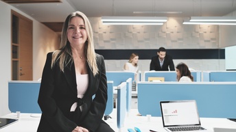 Person mit langen blonden Haaren und Businesskleidung lehnt bei einem Schreibtisch in einem Großraumbüro und blickt freudig in die Kamera, im Hintergrund befinden sich weitere Personen