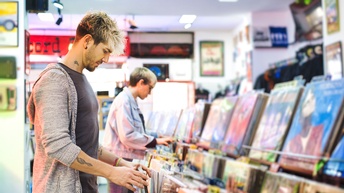 Person steht in Plattenladen vor Regal und blättert Schallplatten durch, im Hintergrund verschwommen weitere Person beim Stöbern im Vinylregal