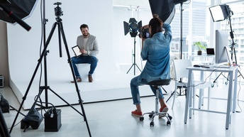 In einem Fotostudio sitzt eine Person vor einem weißen Hintergrund auf einem weißen Hocker. Die Person hält ein Tablet und lächelt. Ihr gegenüber sitzt eine Person, die mit einer Kamera Fotos macht, ringsum stehen Stative mit Lightboxen und Scheinwerfen.