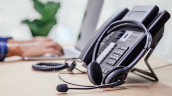 Fokus auf schwarzes Telefongerät mit daranlehnendem Headset, im Hintergrund verschwommen Hände einer Person im Ausschnitt auf Laptop tippend, dahinter Zimmerpflanze