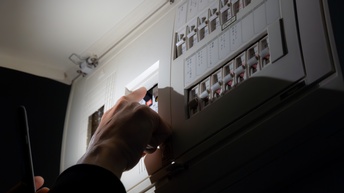 Nahaufnahme der Hand einer Person, die bei einem geöffneten Stromkasten den Fi-Schutzschalter drückt