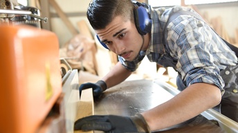 Person mit Ohrenschutz und Schutzbrillen hält Holzbalken auf Werkbank und beugt sich darüber
