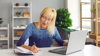 Eine Person mit Brille sitzt am Schreibtisch und schreibt in ihrem Notizblock. Auf dem Tisch liegt ein aufgeklappter Laptop, im Hintergrund stehen Regale und eine große Pflanze.