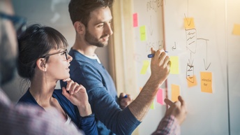 Drei Personen stehen vor Board, eine Person beschriftet Post its