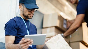 Eine Person mit Kappe, Brille, Bart und Ausweis um den Hals hält in der rechten Hand ein kleines Tablet. In der linken Hand hält sie einen Karton. Im Hintergrund sortiert eine andere Person Kartons
