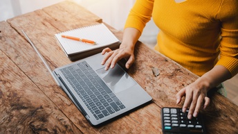Eine Person sitzt an einem Holztisch. Mit der linken Hand tippt sie auf einem Taschenrechner. Mit der rechten Hand bedient sie einen Laptop. Rechts daneben liegt ein Notizblock
