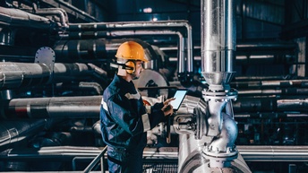 Person in Arbeitskleidung mit orangem Helm und Gehörschutz in Seitenansicht steht in Industrieanlage vor großen Metallrohren und hält Tablet in Händen