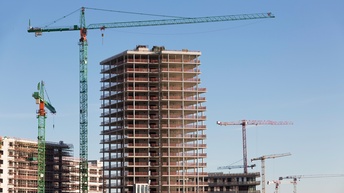 Mehrere Rohbauten von hohen Gebäuden mit vielen Stockwerken unter blauem Himmel. Zwischen den Gebäuden stehen versetzt viele Kräne.