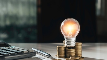 Auf einer Tischplatte sind ein Taschenrechner, ein Stift sowie Münzstapel, auf denen eine leuchtende Glühbirne ist