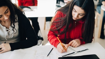 Frontale Aufnahme zweier Personen, die nebeneinander hinter einem Tisch in einem Klassenraum sitzen. Die rechte Person notiert etwas in einem Notizheft. Vor der linken Person ist ein aufgeklapptes Buch