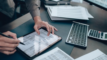Eine Person tippt auf einem Tablet, auf dem eine Tabelle zu sehen ist. Es sind nur ihre Hände zu sehen. Das Tablet liegt auf einem Schreibtisch, auf dem Unterlagen und ein Taschenrechner liegen. 