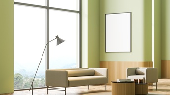 In einem hohen Raum mit einem großen Fenster und hellgrünen Wänden stehen ein helles Sofa und ein Couchsessel. Links vom Sofa ist eine Stehlampe. Vor dem Sofa ist ein runder Teppich, auf dem zwei runde, niedrige Tische sind