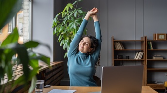 Eine Person in einem bläulichen Oberteil sitzt an einem Tisch. Die Person hat die Augen geschlossen und streckt ihre beiden Arme nach oben. Auf dem Tisch ist ein aufgeklappter Laptop. Im Hintergrund ist ein Regal mit Büchern