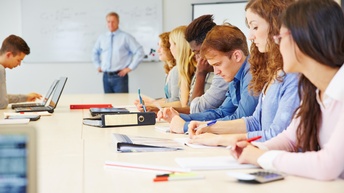 Mehrere Personen sitzen an einem großen Besprechungstisch und vorne steht eine weitere Person bei einer weißen Tafel