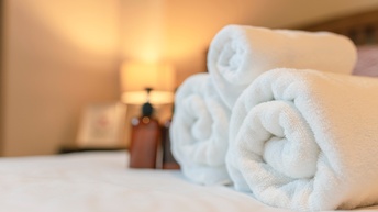 Nahaufnahme dreier eingerollter Handtücher, die auf einem Bett mit weißer Bettwäsche liegen. Daneben stehen braune Plastikspender.