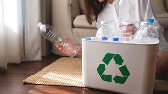 Eine Person sitzt vor einem Sofa auf einem Teppich. Vor der Person ist ein Plastikbehälter, in dem verschiedene Plastikflaschen sind. Auf dem Gefäß sind drei grüne Pfeile gedruckt, die einen Kreis bilden. Die Person hält in jeder Hand eine Plastikfalsche