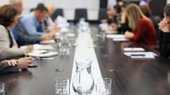 An einem langen Tisch sitzen sich viele Personen gegenüber. Vor den Personen liegen jeweils Unterlagen, in denen die Personen lesen. In der Mitte des Tisches stehen Gläser und gefüllte Wasserkrüge