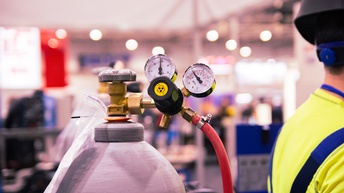 Detailansicht einer Gasflasche mit Druckmessern und Schlauch, im Seitenrand im Ausschnitt Person mit Helm