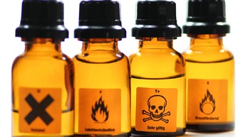 Braune kleine Glasfläschchen mit schwarzen Verschlüssen und Etiketten mit schwarzem X, Flammen und Totenkopf, Fokus auf Letzterem vor weißem Hintergrund