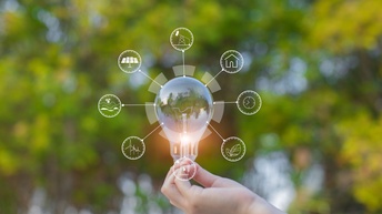 Glühbirne wird von einer Hand in die Höhe gehalten und diverse Icons zum Thema Energiesparen und Nachhaltigkeit strahlen darum, im Hintergrund zeigt sich ein florales Bokeh aus gelb und grünen Farben