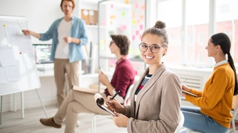 Lächelnde Person mit Brillen sitzt auf Stuhl und hält Stift auf Notizblock, im Hintergrund verschwommen weitere Personen auf Stühlen sitzend auf Person vor Whiteboard stehend blickend