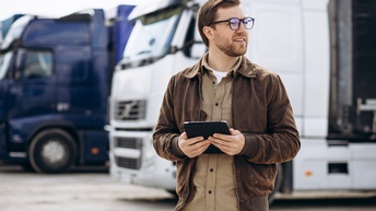 Frontale Aufnahme einer Person mit Brille, kurzen Haaren und Bart, die auf einem Parkplatz im Freien steht, ein Tablet hält und nach rechts blickt. Hinter der Person parken zwei Lastkraftwagen