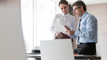 Zwei Personen in Businesskleidung besprechen Unterlagen in einem Büro, eine Person trägt eine Brille sowie einen Bart