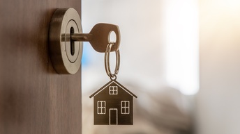 Nahaufnahme eines Schlüssels in einem Türschloss. Am Schlüssel hängt ein Schlüsselanhänger in Form eines Hauses