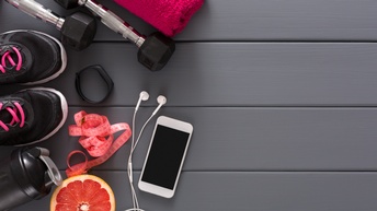 Fitnessschuhe, Hanteln, Handtuch, Getränkeflasche, Sportuhr, Maßband sowie eine Grapefruitscheibe, Smartphone und Kopfhörer liegen auf einem grauen Dielenboden