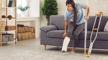 Person mit eingegipsten Bein hält sich an Krücke fest und versucht sich auf grauer Couch niederzusetzen, an Couch weitere Krücke lehnend, im Hintergrund verschwommen Regal mit Büchern und Schachteln sowie Zimmerpflanzen