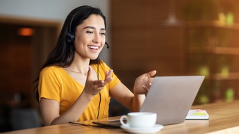 Person mit Headset und gelbem T-Shirt sitzt freudig vor einem Laptop und hält eine Konferenz, daneben steht eine Tasse sowie liegt ein Notizbuch auf dem Holztisch in einem Café