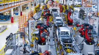 Top-Shot einer großen Fabrik, in der die Hülle von grauen Autos sind. Links und rechts der Autos sind große, rote Roboterarme, die auf grauen, hohen Plattformen stehen.