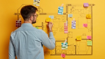 Rückenansicht einer Person, die vor einer gelben Wand steht. Auf der Wand ist ein Grundriss eines Stockwerks eines Gebäudes aufgezeichnet. Im Grundriss sind verschiedene Räume und Möbelstücke eingezeichnet. Daneben hängen Farbpaletten