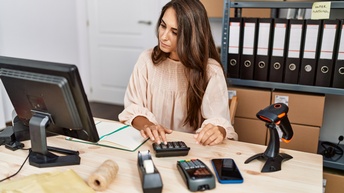 Person sitzt an einem Schreibtisch und blickt auf ihren Bildschirm, während sie einen Taschenrechner bedient, im Hintergrund steht ein Regal mit Ordnern und Kartonagen sowie stehen am Schreibtisch noch ein Scan- und ein digitales Kartenbezahlgerät, ein Smartphone und Verpackungsmaterialien