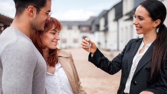 Lächelnde Person reicht zwei aneinander geschmiegten ebenfalls lächelnden Personen Schlüssel, im Hintergrund verschwommen Häuserreihen