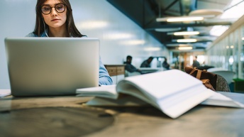 Person mit Brillen und Kopfhörern in den Ohren sitzt in Lesesaal und blickt auf aufgeklapptes Notebook vor ihr, am Tisch im Vordergrund verschwommen Bücher liegend