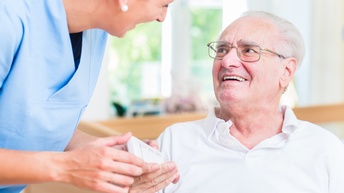 Nahaufnahme einer sitzenden Person mit kurzen, grauen Haaren und Brille, die zu einer links neben ihr stehenden Person blickt, die sie ebenfalls anblickt. Die linke Person trägt blaue Kleidung und beugt sich leicht nach vorne