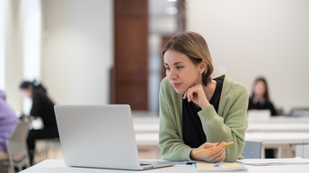 Person mit kurzen Haaren sitzt an einem Tisch in einem Raum und blickt auf einen Laptopbildschirm. In der rechten Hand hält sie einen. Daneben liegen weitere Unterlagen auf dem Tisch. Im Hintergrund sind weitere Tische, an denen Leute sitzen