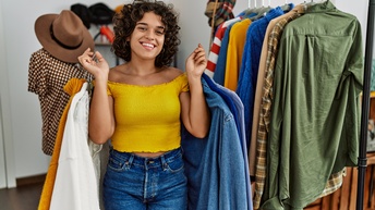 Porträt einer lächelnden Person, die in einem Geschäft steht und in beiden Händen Kleiderhaken mit Kleidungsstücken hält. Neben ihr steht ein Kleiderständer mit weiteren Kleidungsstücken
