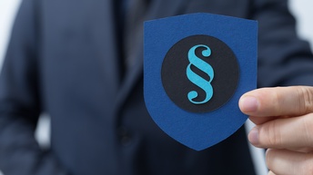 Nahaufnahme der linken Hand einer Person, ansonsten ist nur der Oberkörper ohne Gesicht verschwommen zu sehen. Sie hält ein blaues Papier in Wappenform in der Hand, auf dem ein schwarzer Kreis klebt. Auf diesem klebt ein türkises Paragraphen-Zeichen. 